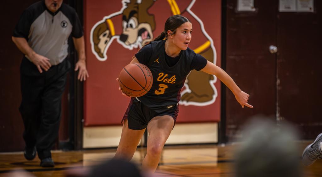 image of Minneapolis Roosevelt High School girls basketball guard dribbling the ball up the court during a game. The Teddy illustrated mascot logo, printed on a wall cushion under the basket, is visible directly behind her