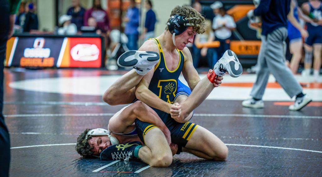 Image of the Minneapolis Roosevelt High School Teddy Wrestler during a match. He’s kneeling on his knees sitting on top of his opponent wrapping him up.
