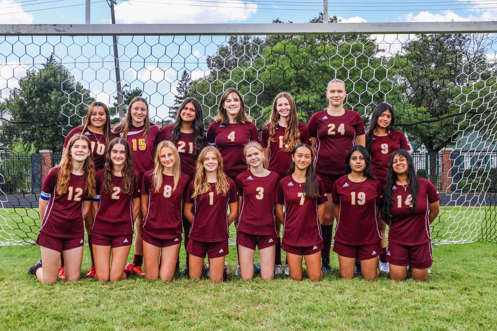 Image of Minneapolis Roosevelt High School’s girls JV soccer team of 2025 grouped together with their coach for a team photo on the field at Al Gowans Stadium. Superimposed over the photo in the lower right corner is the Soccer program logo.